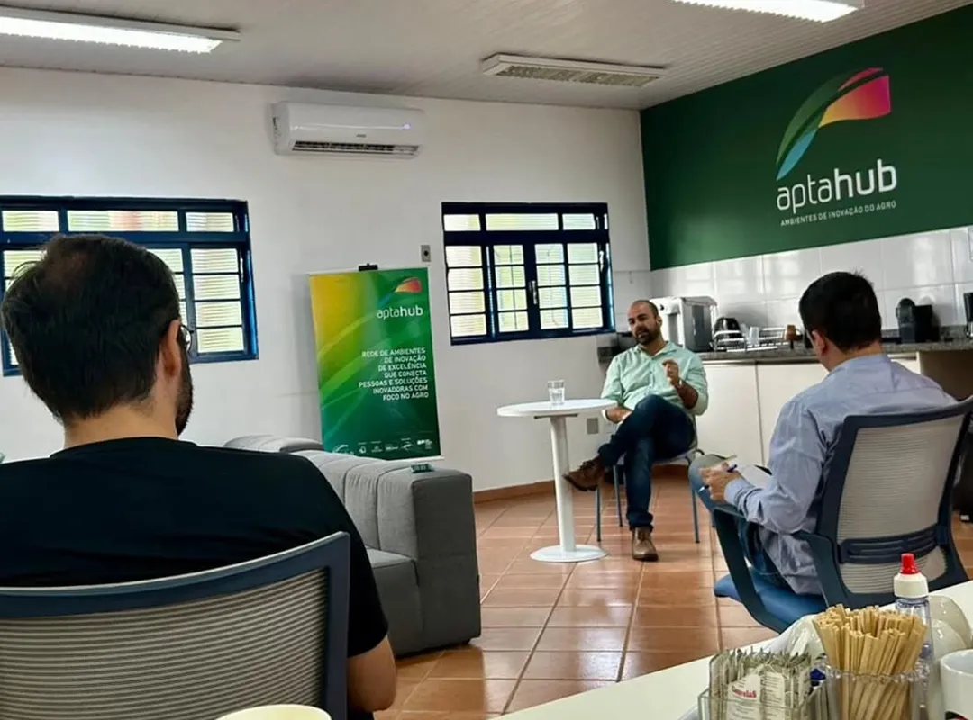 ambiente interno; há três pessoas na imagem, sendo que uma delas está falando. Ao fundo, uma placa com texto AptaHub.